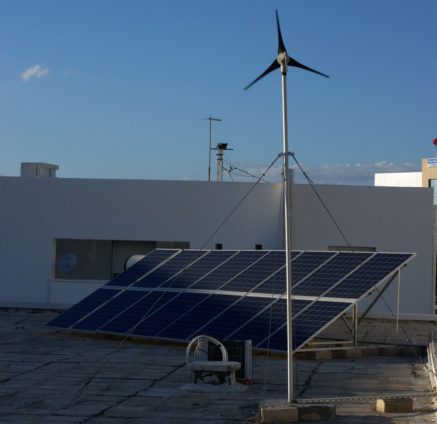 Sur un toit-terrasse blanc, une rangée de panneaux solaires et une petite éolienne sur un mât haubané.